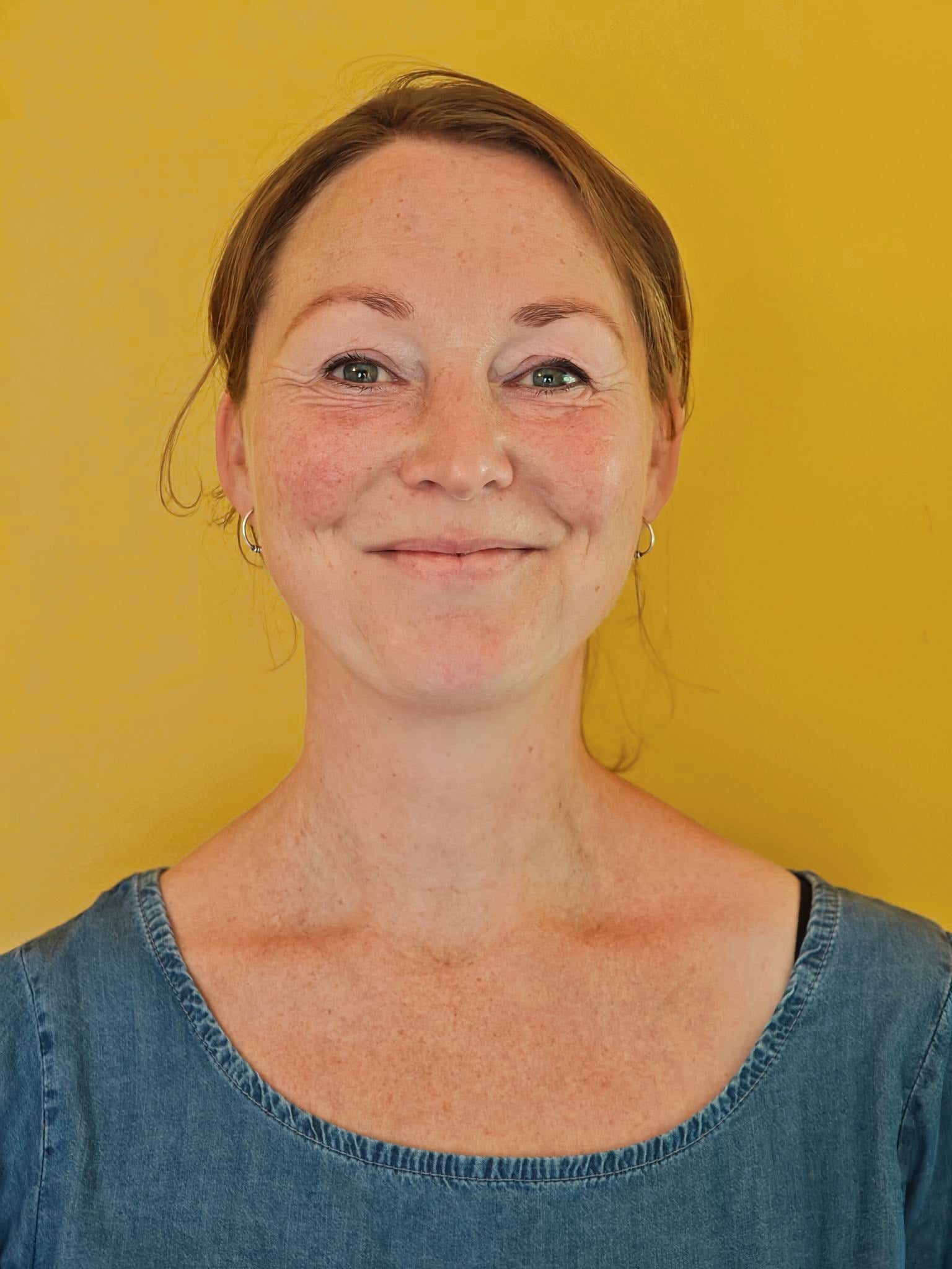 Moira kijkt in de camera en glimlacht, tegen een warmgele achtergrond